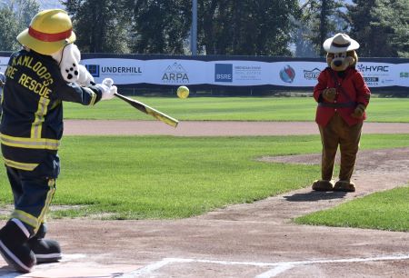 Sparky, la mascotte du service d’incendie et de sauvetage de Kamloops (un dalmatien portant un uniforme de pompier), prend son élan pour frapper une balle molle lancée par le sergent Ours prudent, la mascotte de la GRC de Kamloops (un ours portant une tunique rouge et un chapeau de la GRC). 