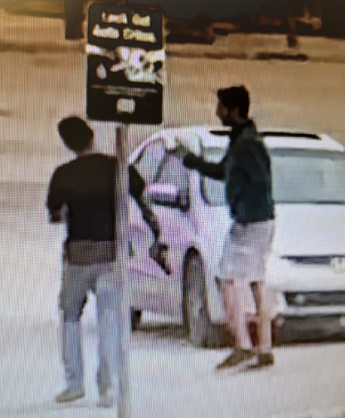 Two South Asian men with dark tops, standing in front of a silver car with one holding a potential handgun